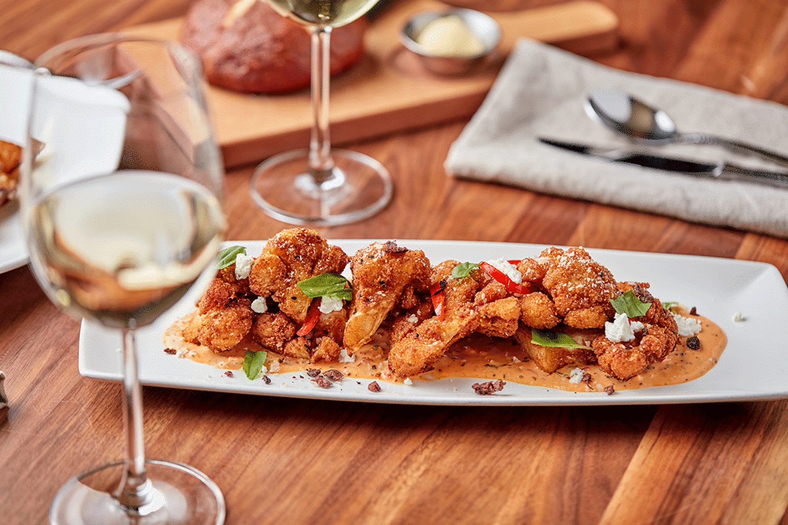Plated crispy fried seafood or chicken bites on creamy sauce, garnished with herbs and chili, on a wooden table with wine glasses and napkins