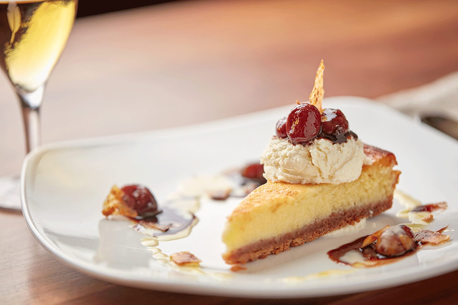 Slice of cheesecake topped with vanilla ice cream and cherries on a white plate, with caramel sauce and a blurred champagne flute in the background