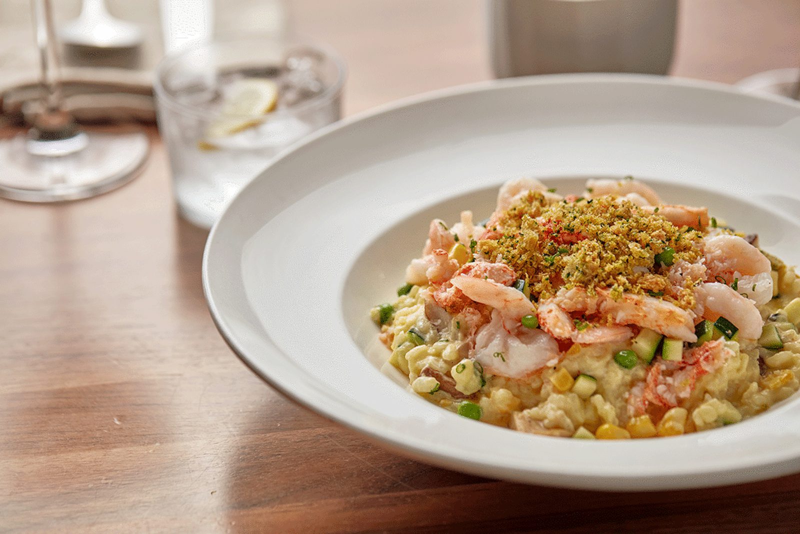 White bowl of shrimp risotto topped with breadcrumbs and herbs on a wooden table, with a glass of water blurred in the background