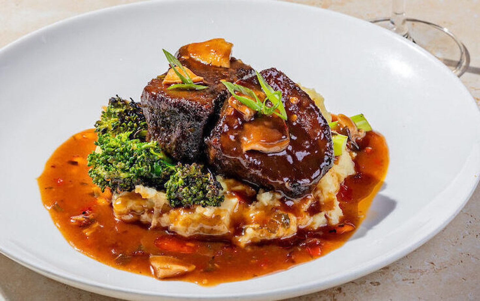Braised short ribs in brown sauce over mashed potatoes with roasted broccoli, sliced mushrooms and green onion garnish