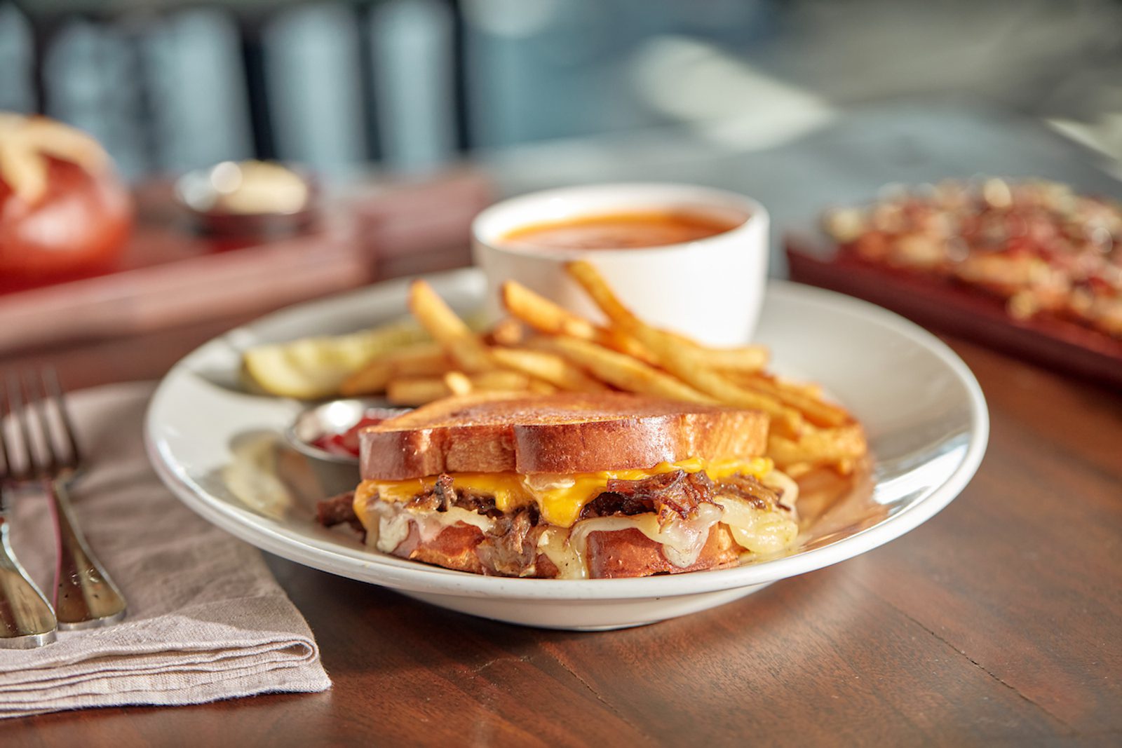 Grilled sandwich with melted cheese, fries, and a cup of dipping sauce on a white plate on a wooden table, with napkin and utensils nearby