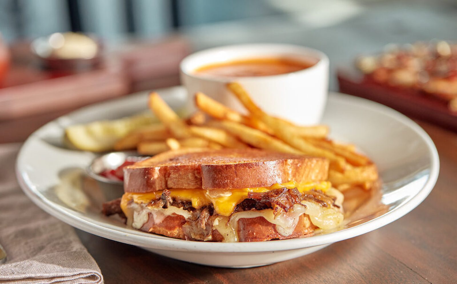 Grilled cheese sandwich with melted cheese on a plate with fries, pickle slices, ketchup, and a cup of tomato soup on a wooden table