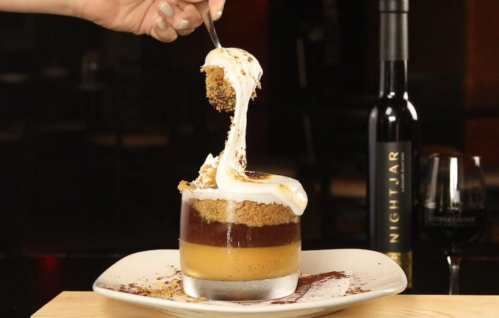 Hand lifting a spoonful of layered dessert with cream on top, served in a glass on a plate, with a wine bottle and glass in the background