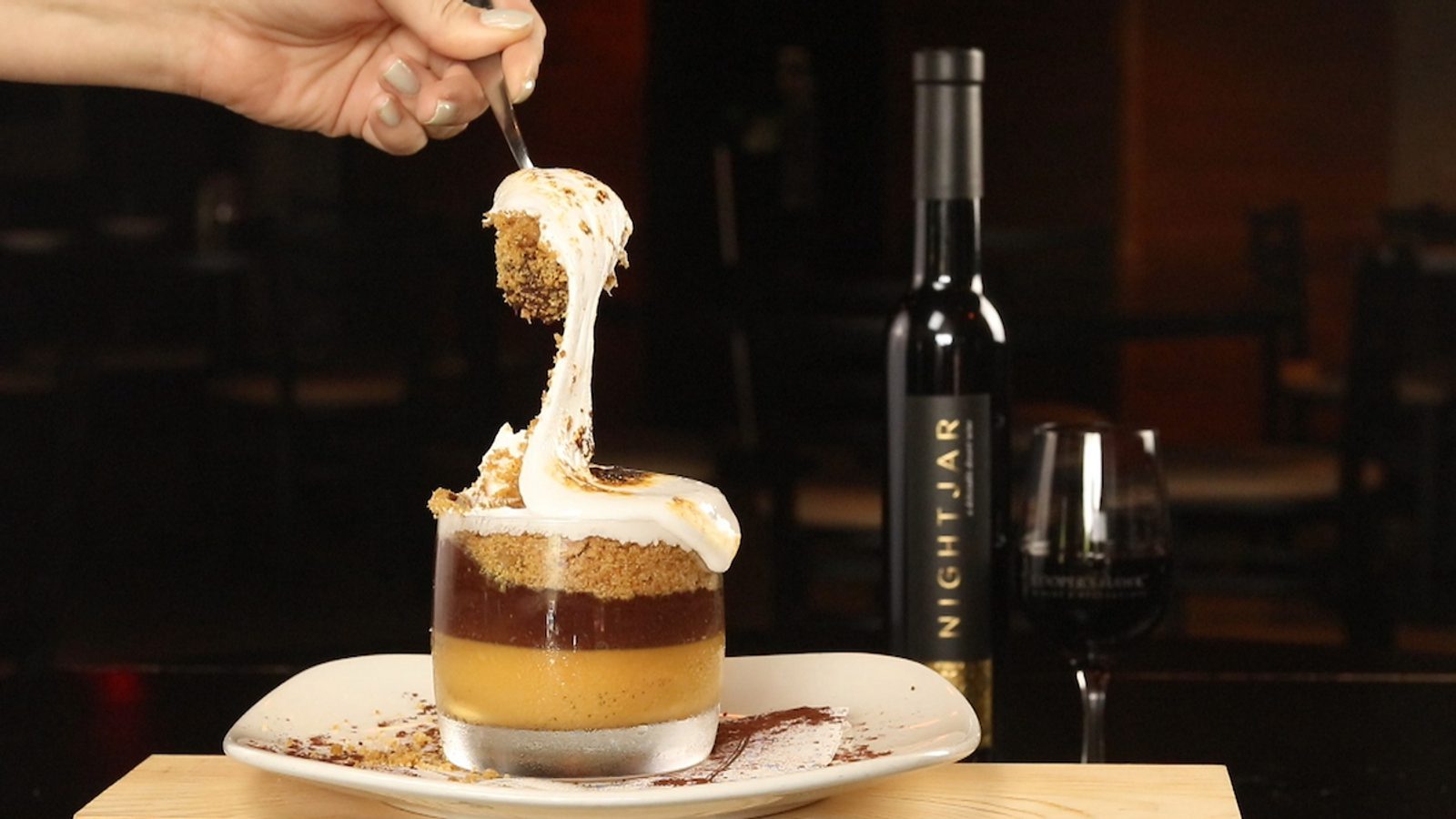 Hand lifting a spoonful of layered dessert with cream on a white plate, with a wine bottle and glass blurred in the background on a dark table