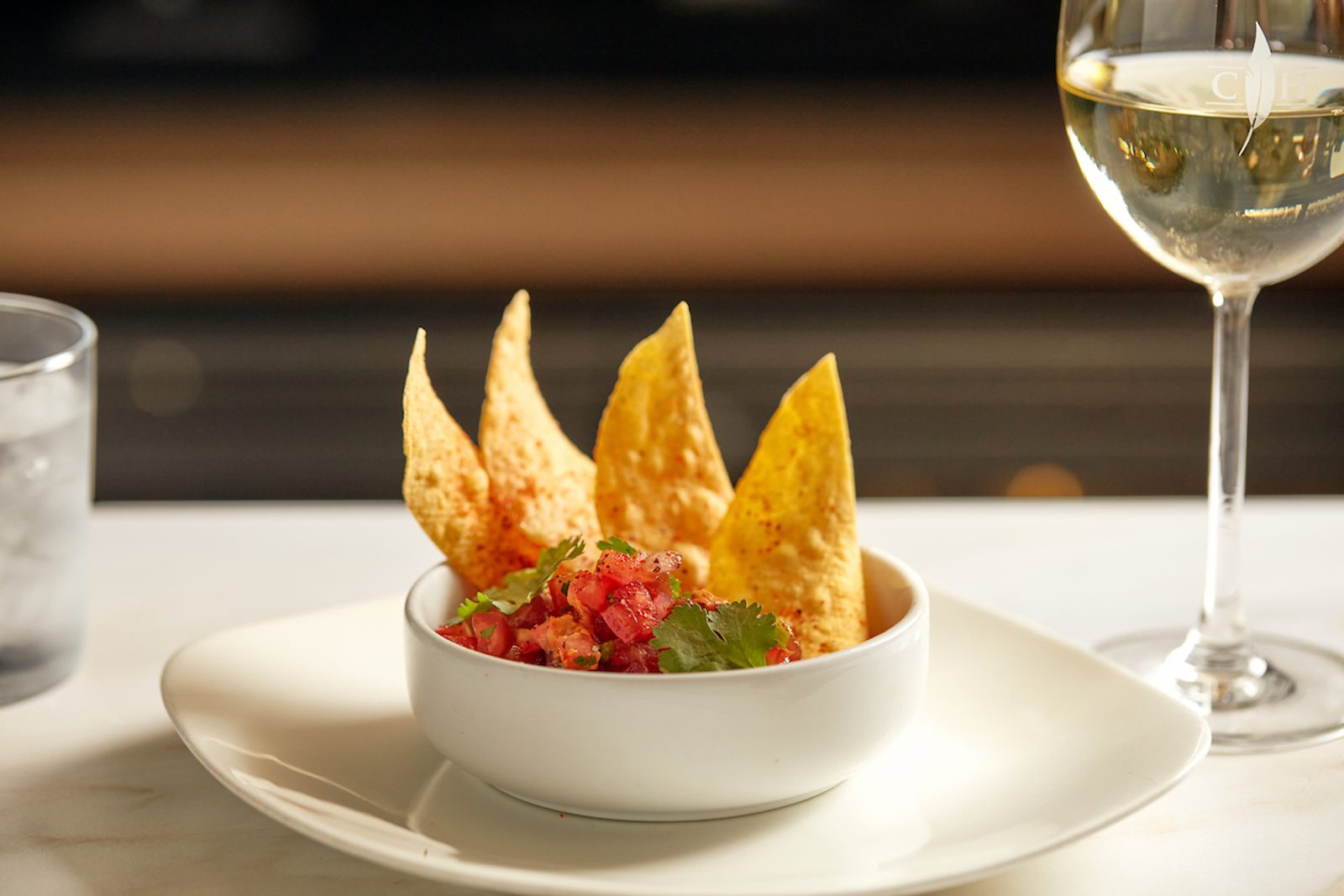 Bowl of salsa topped with cilantro, served with tortilla chips on a white plate beside a glass of white wine on a restaurant table