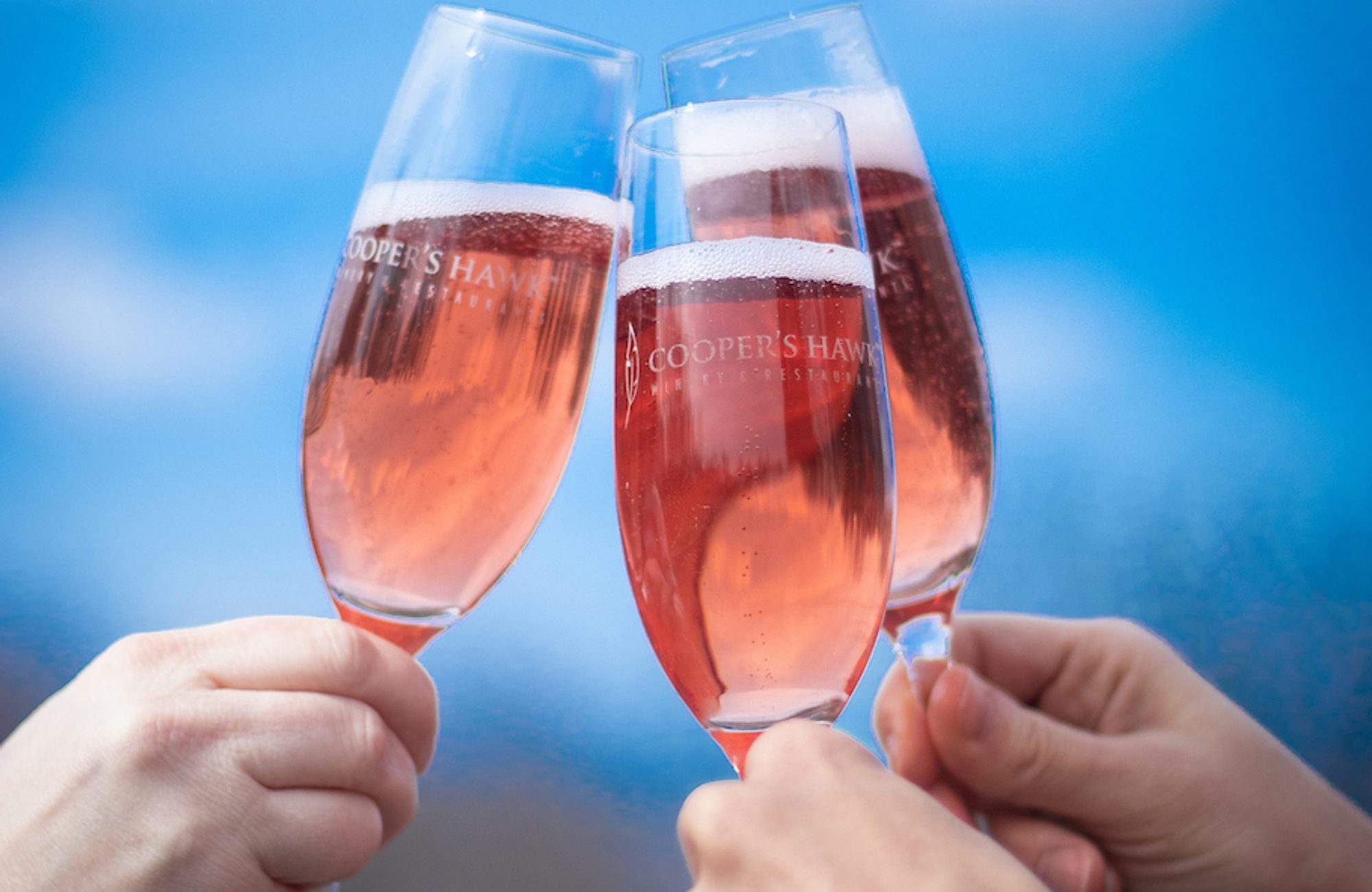 Three hands clinking champagne flutes of rosé sparkling wine, glasses labeled COOPER'S HAWK