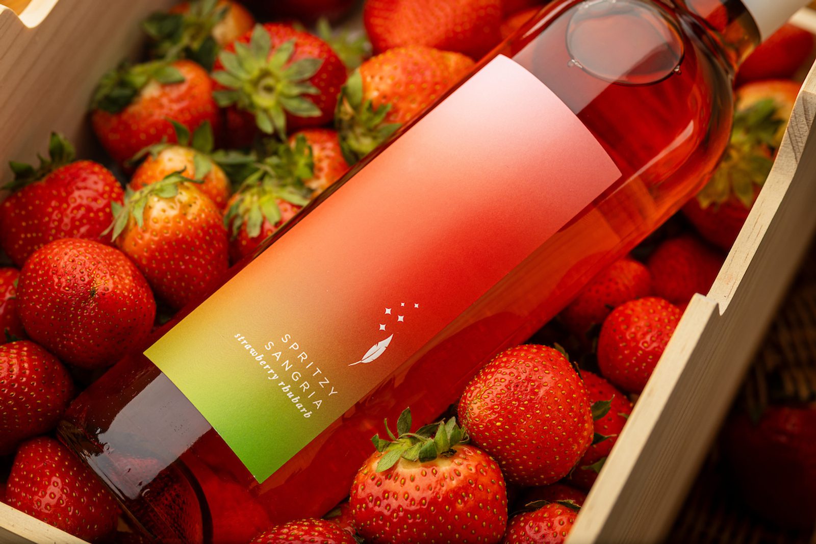 Bottle labeled SPRITZY SANGRIA with red-to-green gradient label resting on fresh strawberries in a wooden crate