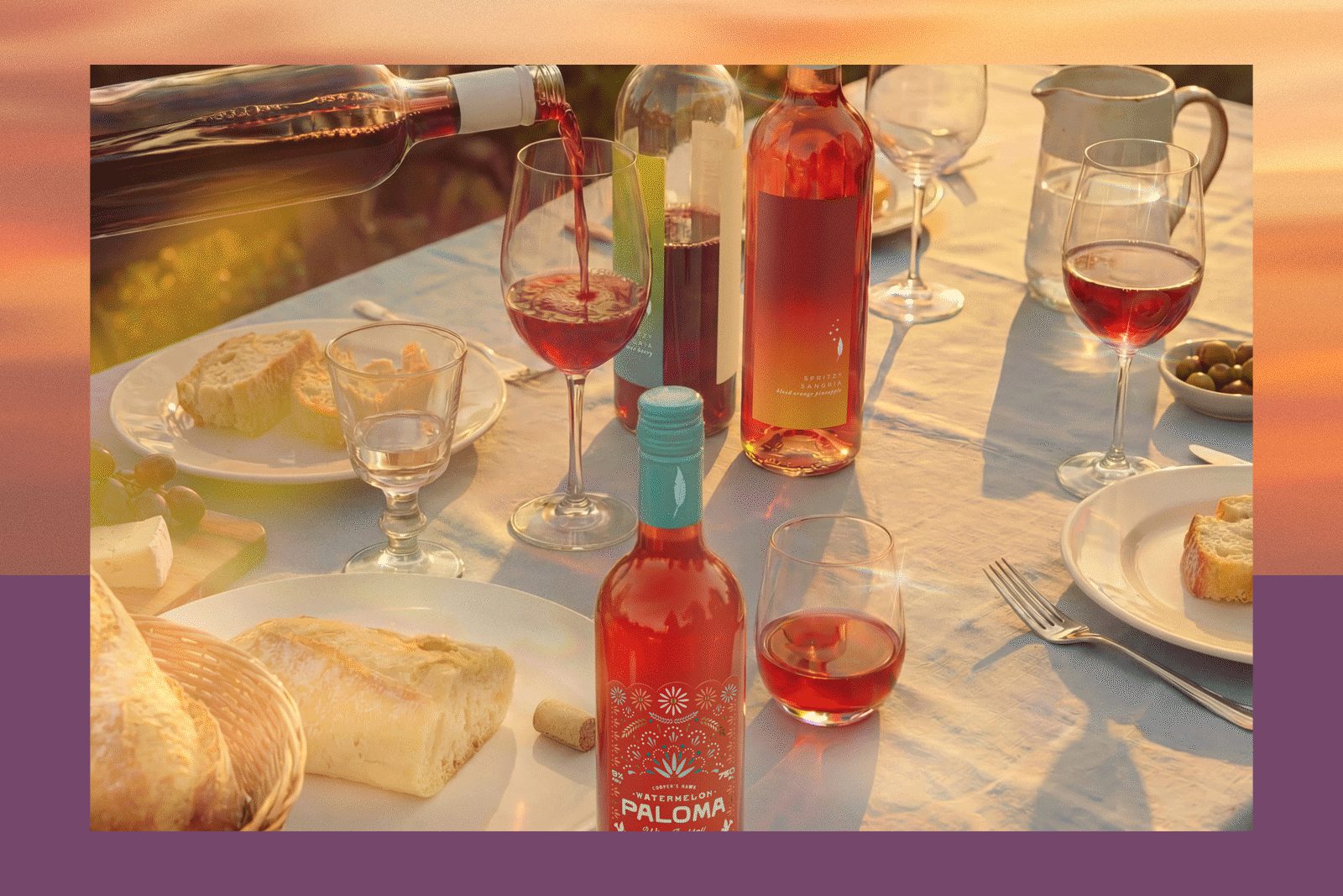 Table setting with rosé bottles (label reads 'Watermelon Paloma'), wine being poured into glasses, bread, cheese, and olives.