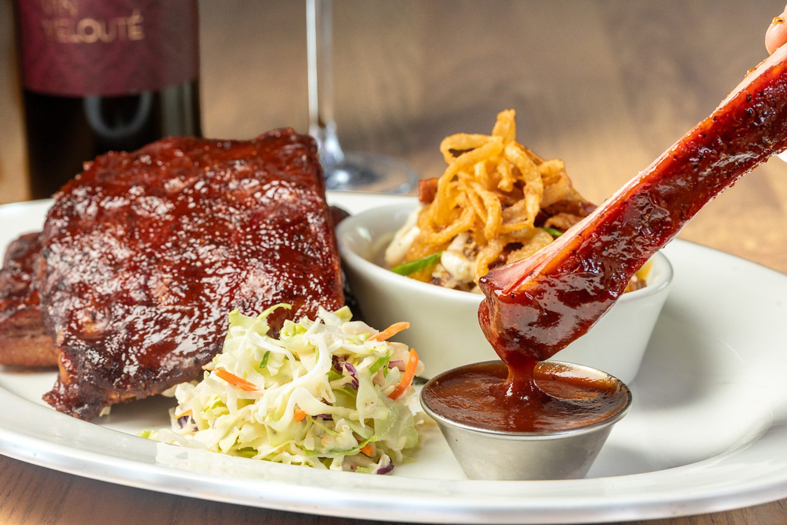 Barbecue-glazed pork rib being dipped into sauce on a plate with coleslaw and a side topped with crispy fried onions