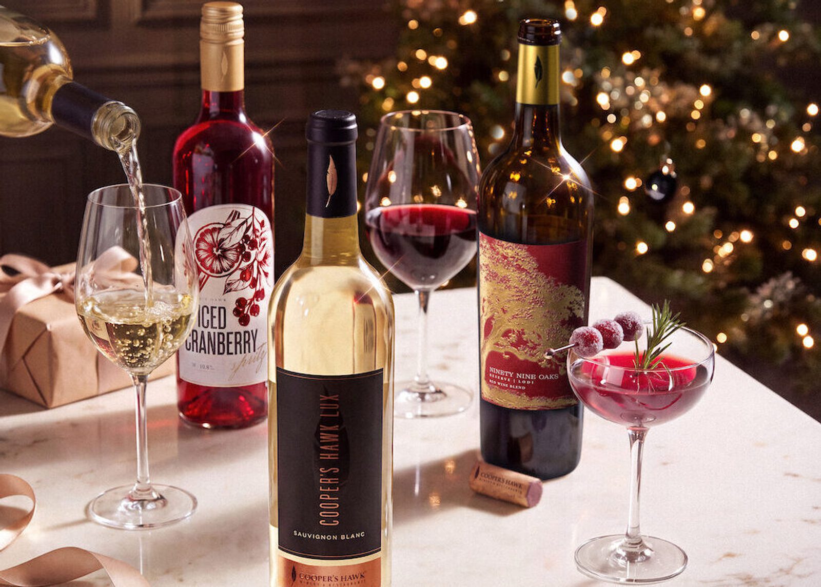 Wine bottles and filled glasses on a festive table, including red, white, and cranberry wine cocktails, with gift wrap and a softly lit Christmas tree in the background