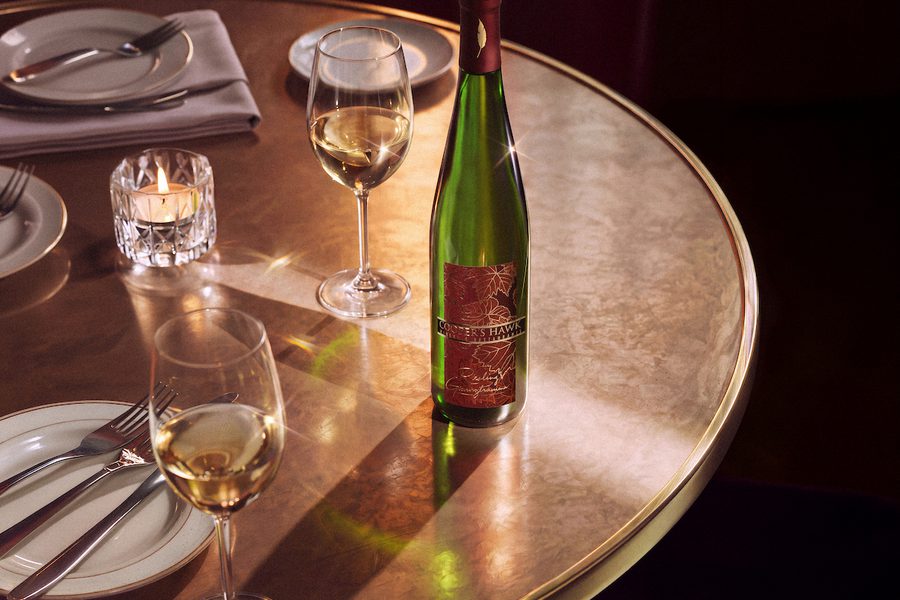 Elegant restaurant table setting with a green wine bottle, two filled wine glasses, plates with silverware, and a small lit candle on a round table