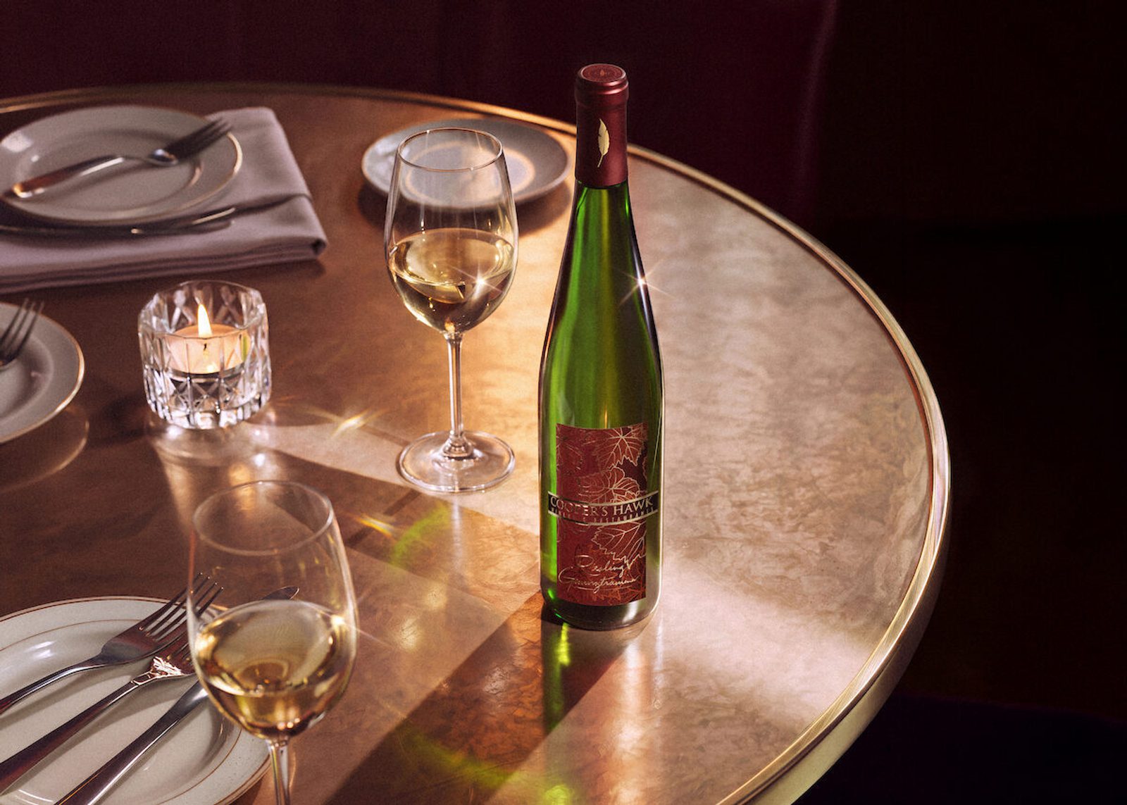 Elegant restaurant table set with a wine bottle, two glasses of white wine, a lit candle, plates, and folded napkins on a polished round tabletop
