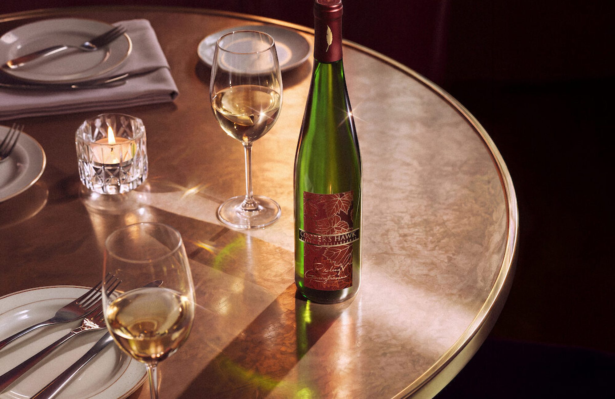 Elegant restaurant table set with a wine bottle, two glasses of white wine, a lit candle, plates, and folded napkins on a polished round tabletop
