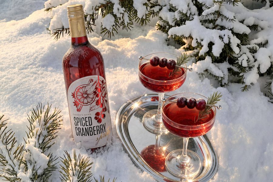 Bottle of spiced cranberry drink beside two red cocktails on a silver tray in snow, garnished with cranberries and rosemary, framed by snowy evergreen branches