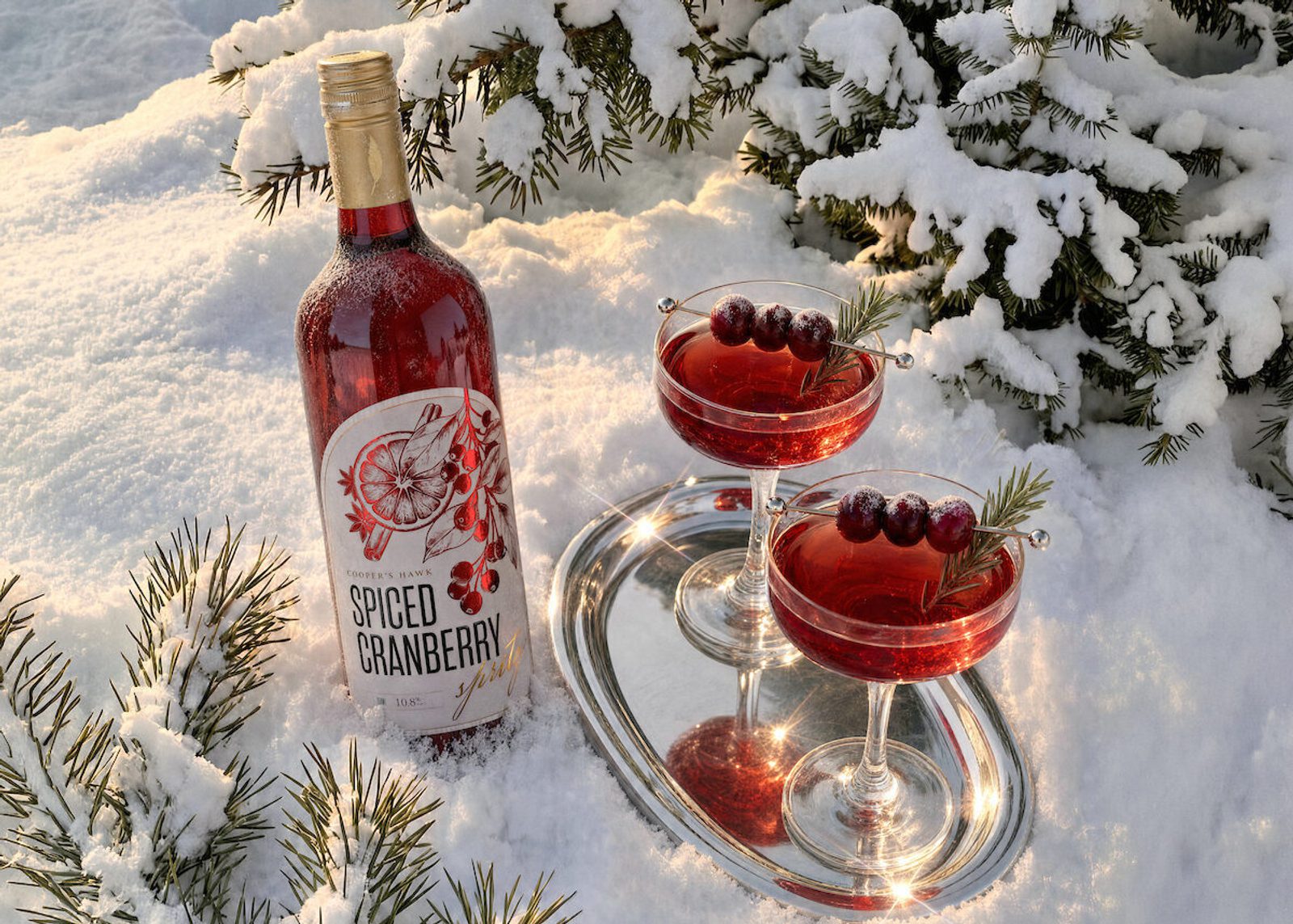 Bottle of spiced cranberry wine beside two garnished red cocktails on a silver tray in fresh snow, with snow-covered pine branches in the background