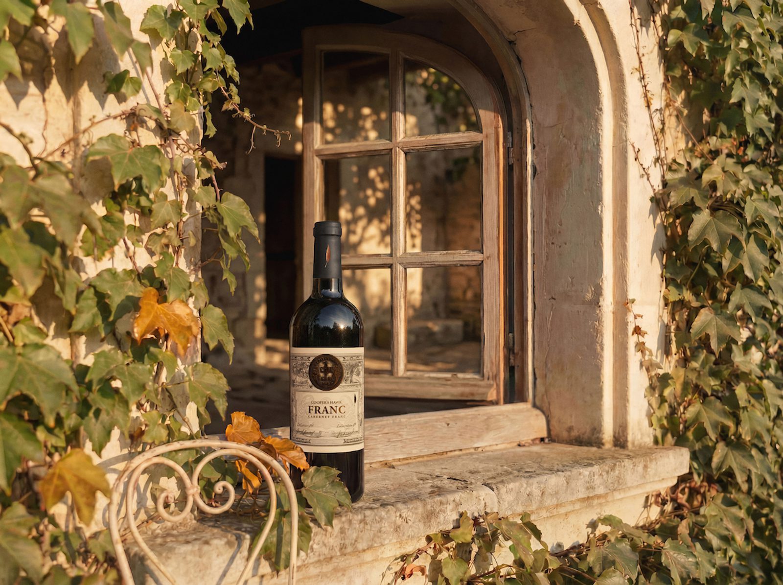 Wine bottle on a rustic stone windowsill beside an arched wooden window, framed by ivy-covered walls in warm sunlight.