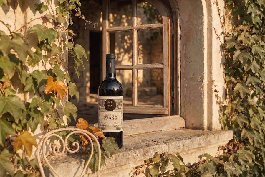 Wine bottle on a rustic stone windowsill beside an arched wooden window, framed by ivy-covered walls in warm sunlight.