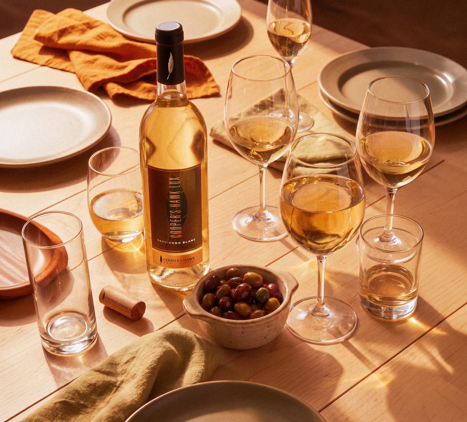 Wine bottle and several filled glasses on a sunlit table with plates, napkins, a bowl of olives, and warm golden afternoon shadows