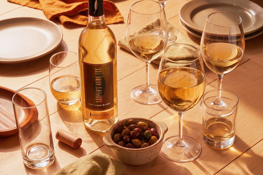 Wine bottle and several filled glasses on a sunlit table with plates, napkins, a bowl of olives, and warm golden afternoon shadows