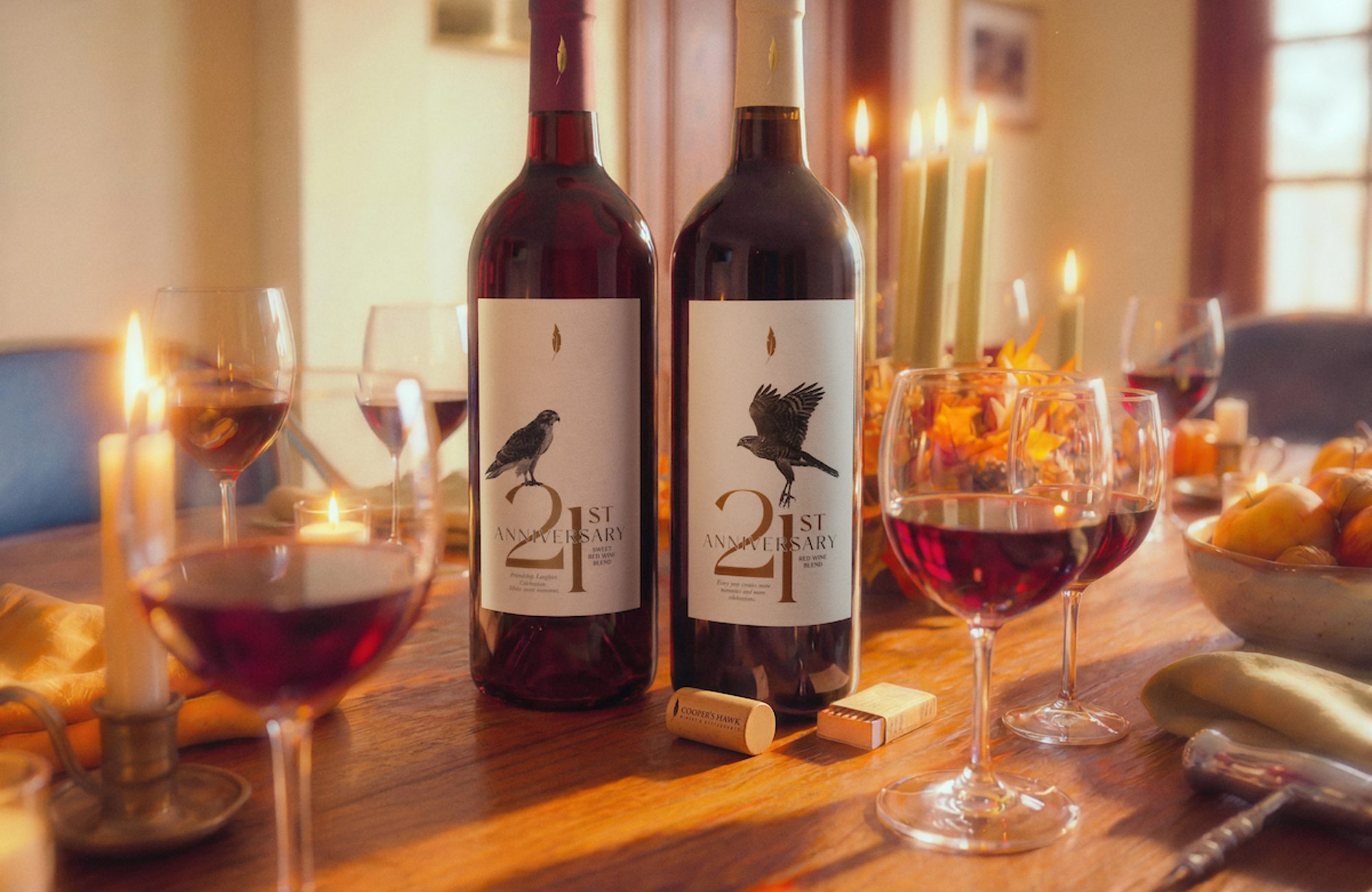 Two labeled wine bottles on a candlelit table with several filled wine glasses, fruit, corks, and warm sunlight streaming through a dining room.
