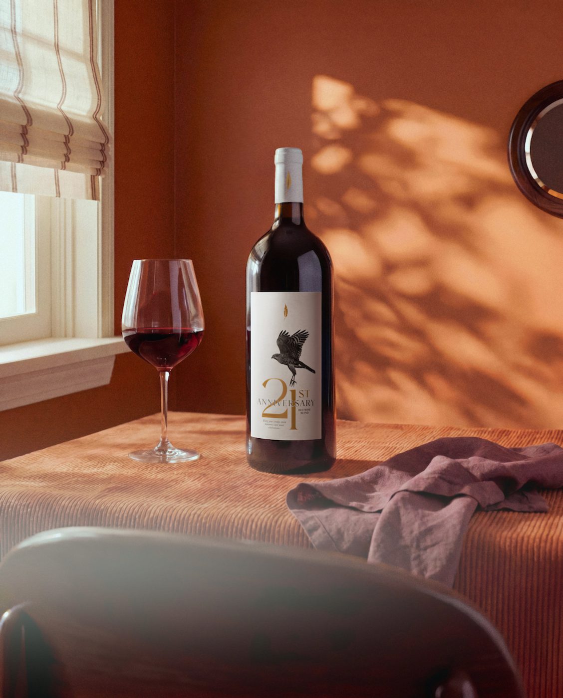 Wine bottle and half-full red wine glass on a sunlit table by a window, with warm shadows on the wall and a crumpled cloth nearby