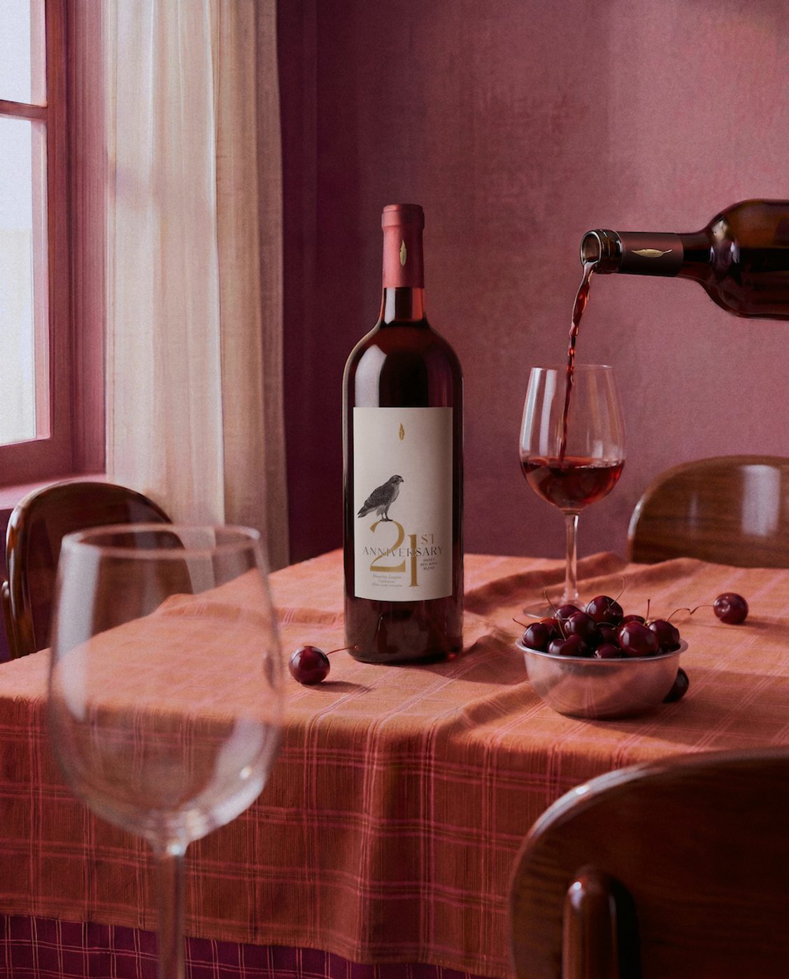 Wine being poured into a glass at a table with a bottle labeled “1st Anniversary,” a bowl of cherries, and a pink-toned dining room with chairs and curtains