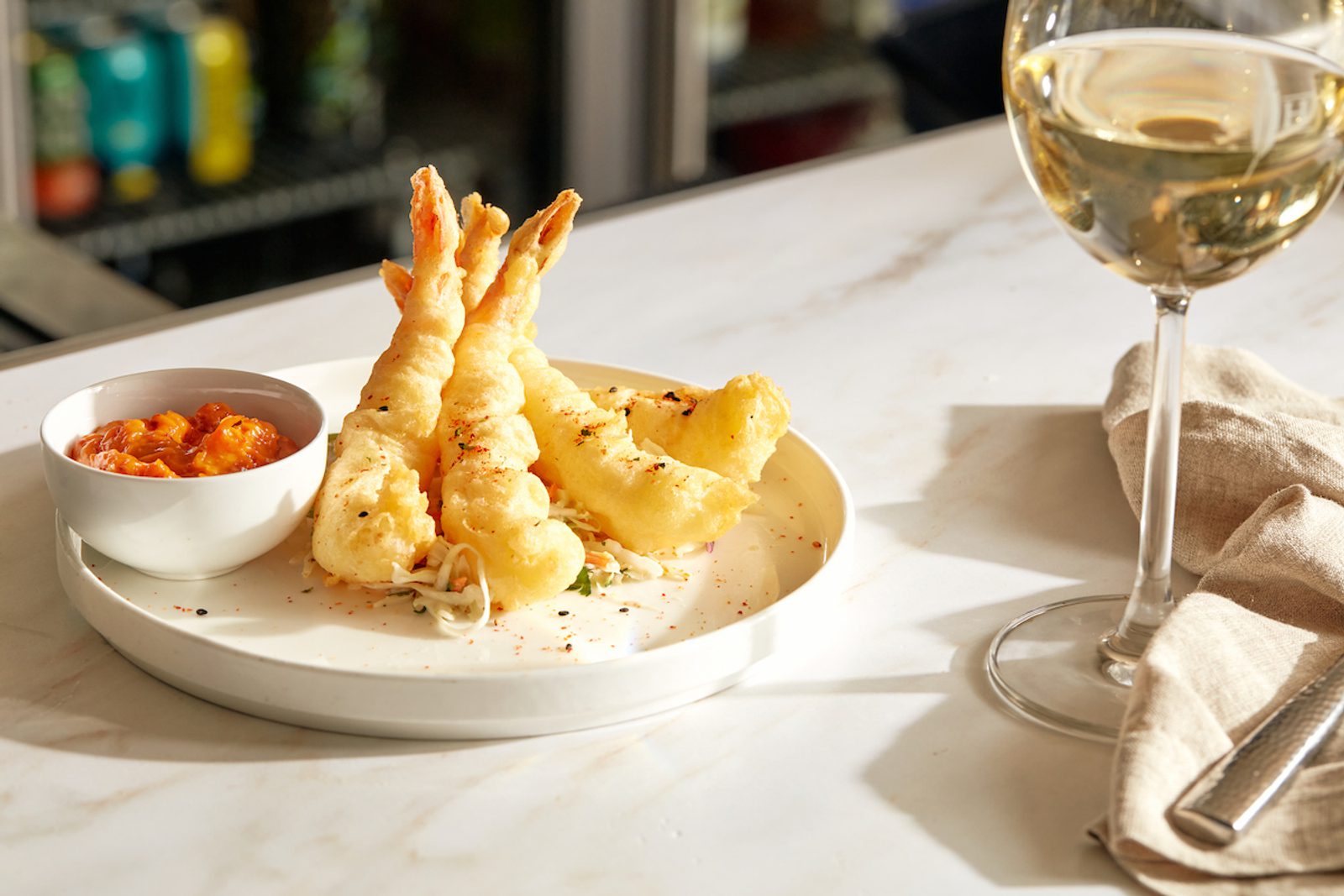 Plate of crispy fried shrimp with dipping sauce beside a glass of white wine on a sunlit table