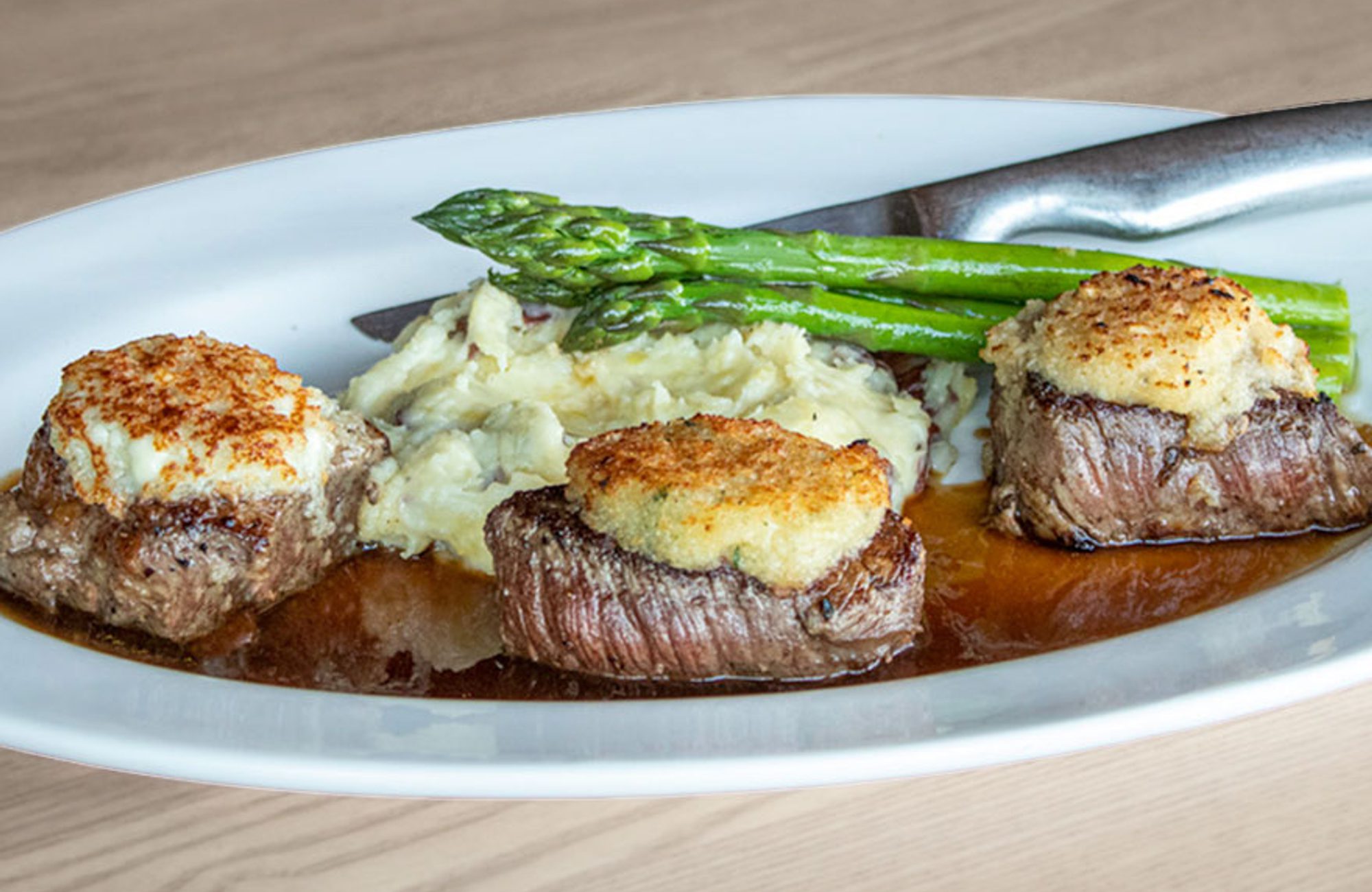 Three beef medallions topped with crab crusts, mashed potatoes, asparagus spears, and brown sauce on a white plate