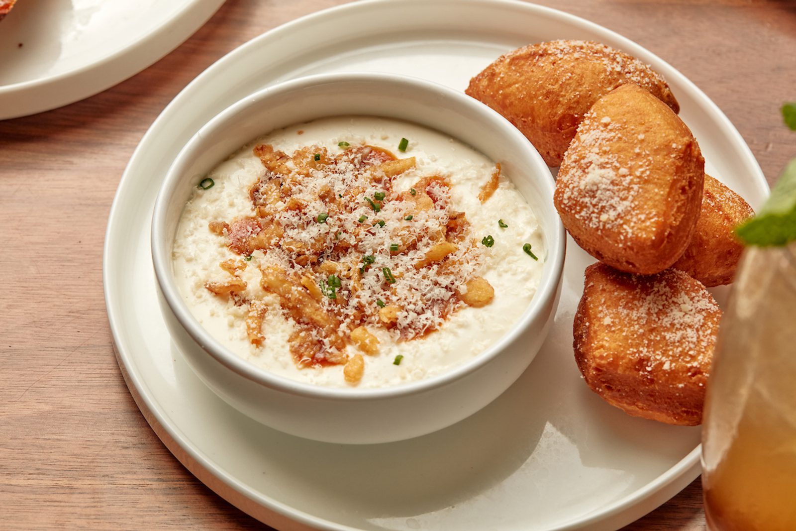 Bowl of creamy dip topped with crispy bacon and cheese, served on a plate with three golden fried bread pieces on a wooden table