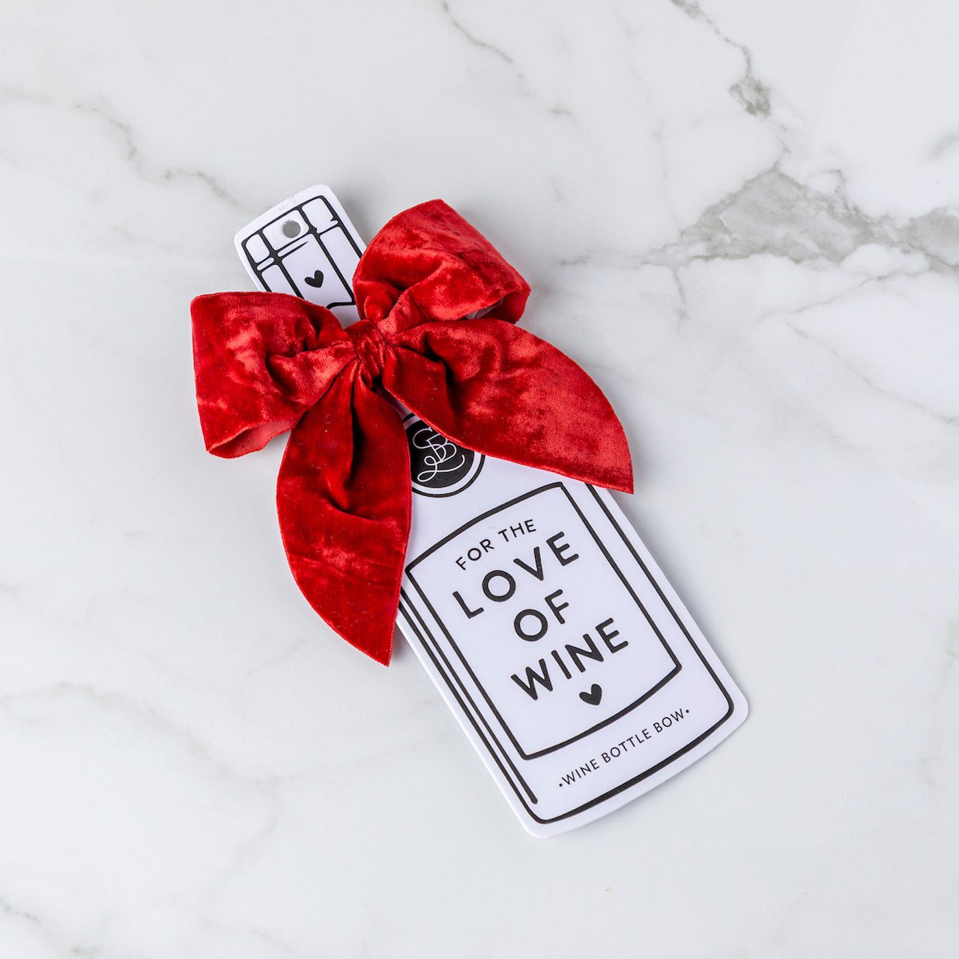 Red velvet bow attached to a wine bottle-shaped gift tag that reads “For the Love of Wine” on a white marble background