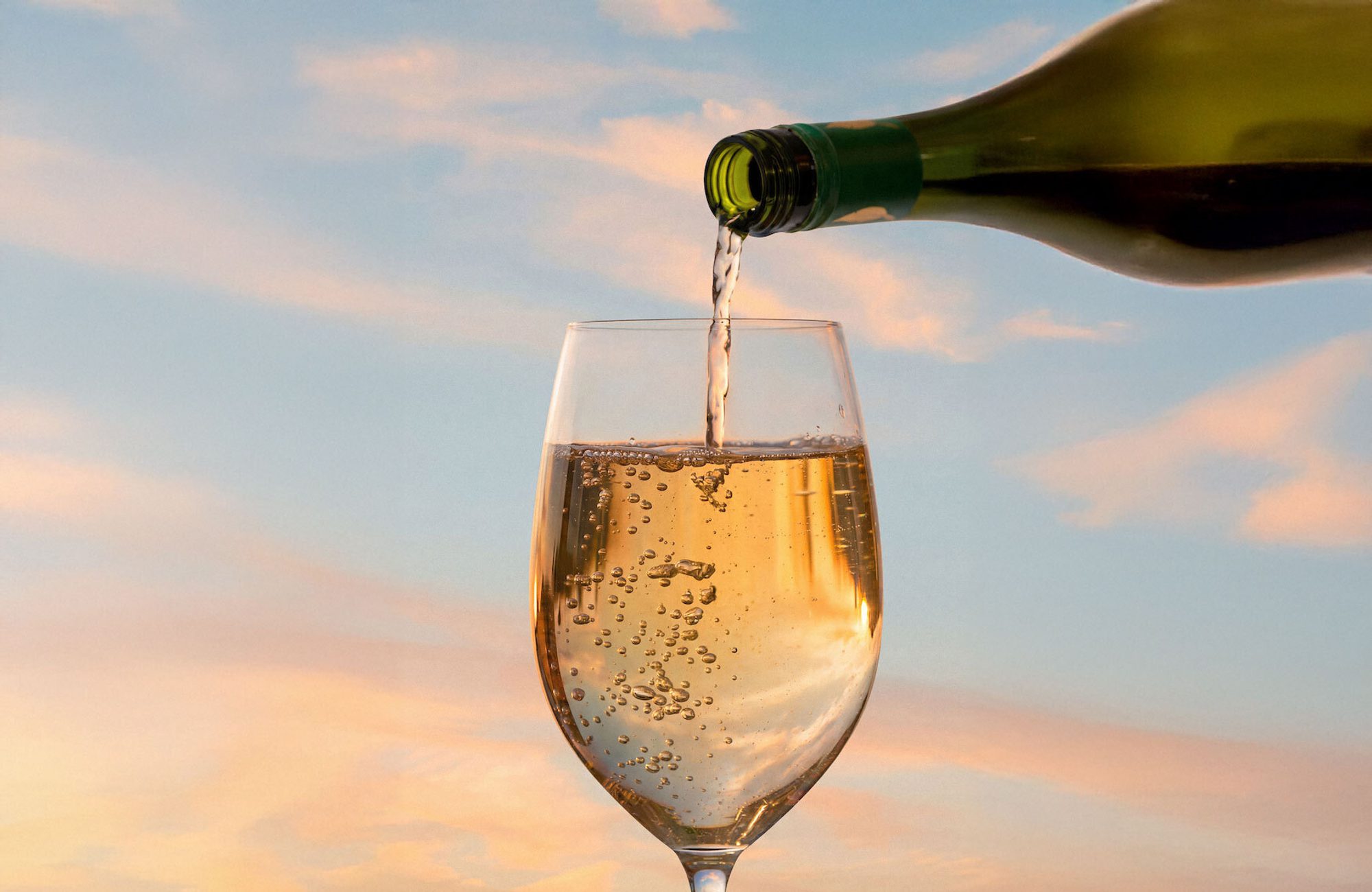 Green bottle pouring sparkling wine into a glass; golden bubbles against a pastel sky