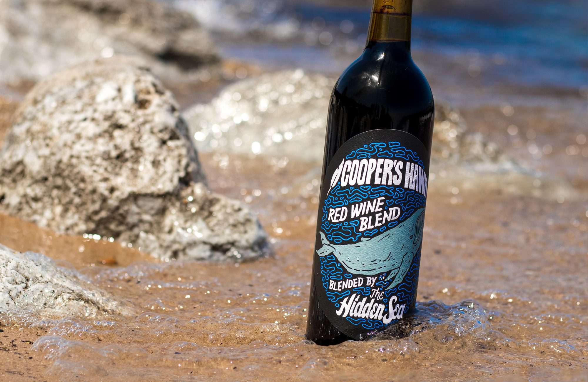 A bottle of wine sitting in the sand next to the ocean.