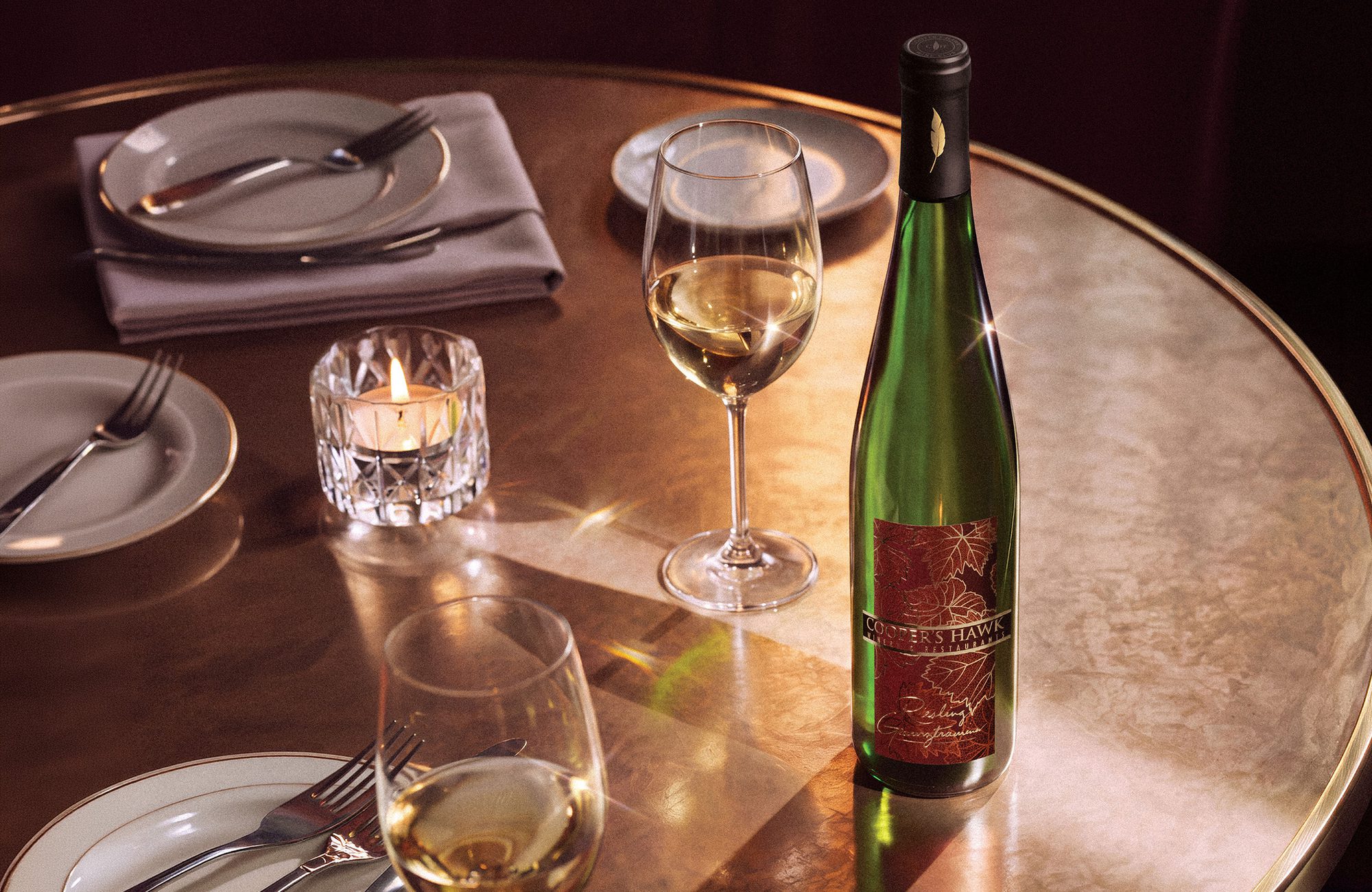 Elegant dining table set with plates, napkins, cutlery, a lit candle, two wine glasses, and a bottle of white wine on a copper-toned surface
