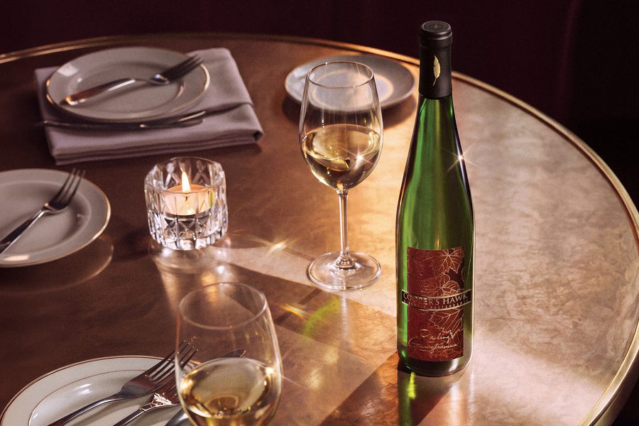 Elegant restaurant table setting with white plates, folded napkins, lit candle, two wine glasses, and a green bottle of white wine on a copper-toned table