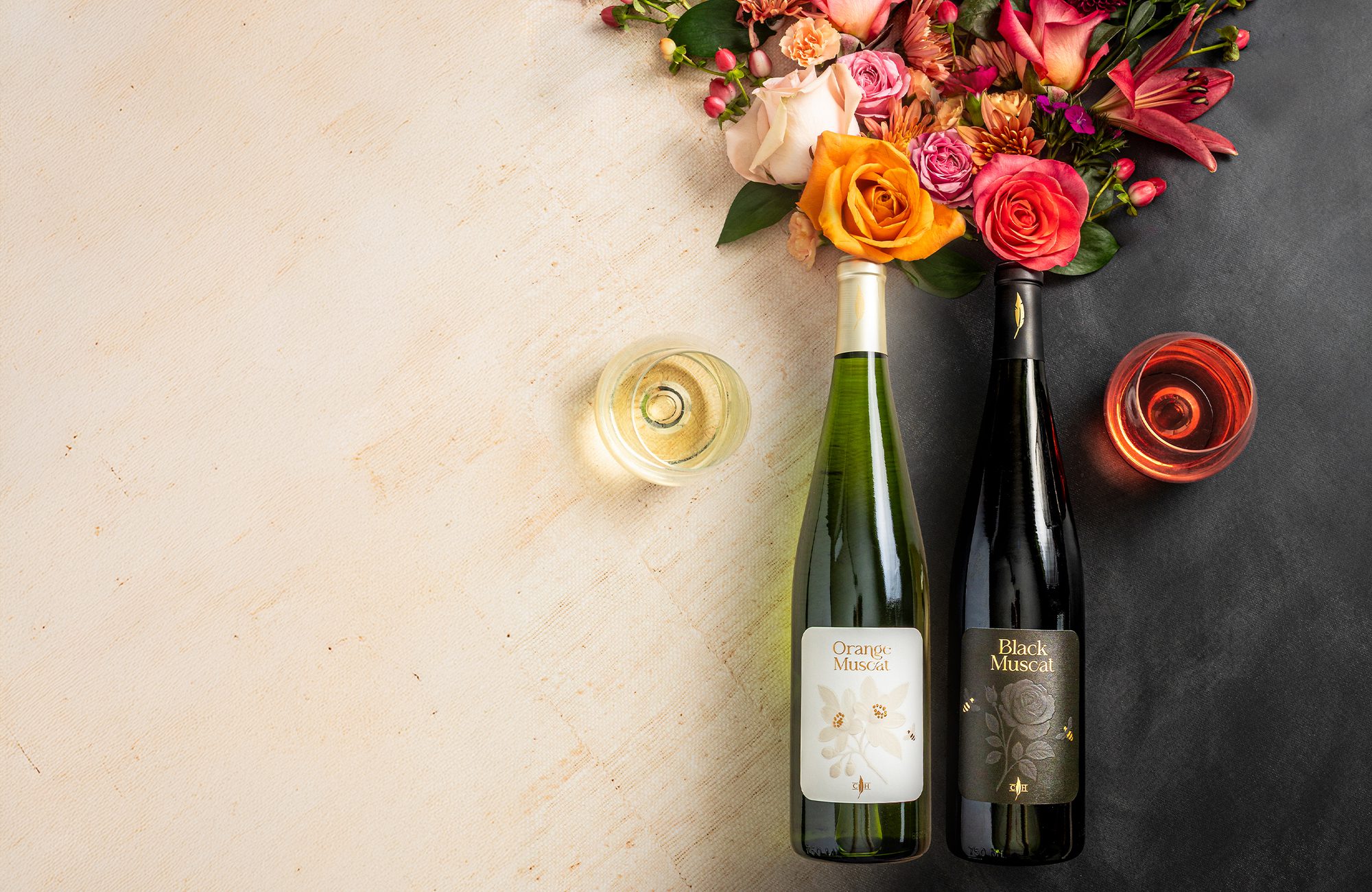 Two bottles labeled Orange Muscat (green) and Black Muscat (dark) with two wine glasses and a floral bouquet on split beige and black background