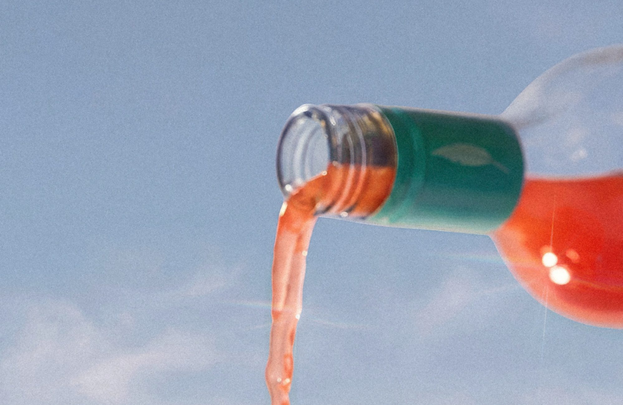 Bottle pouring a pink-red drink into an ice-filled glass against a clear blue sky
