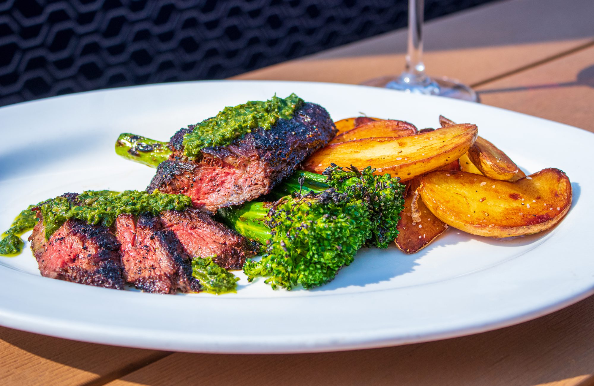 Sliced grilled steak topped with chimichurri, served with broccolini and golden potato wedges on a white plate outdoors