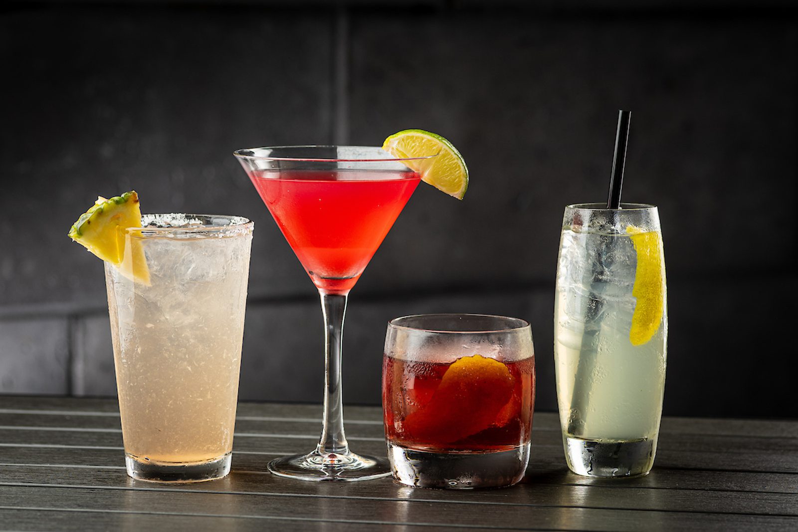 Four colorful cocktails on a dark table: a pineapple-garnished highball, red martini with lime, amber old fashioned, and lemon spritz with straw