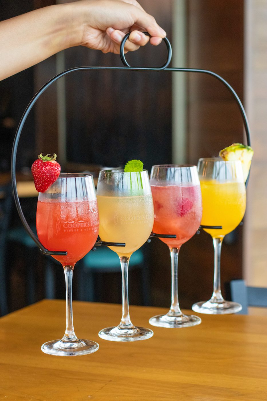 Hand holding a metal carrier with four colorful wine glasses of fruity cocktails, each garnished with fruit, on a wooden table indoors