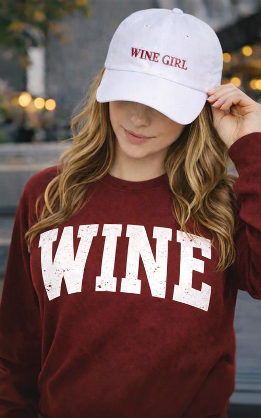 Person adjusting white cap that reads 'WINE GIRL' and wearing maroon sweatshirt that reads 'WINE'