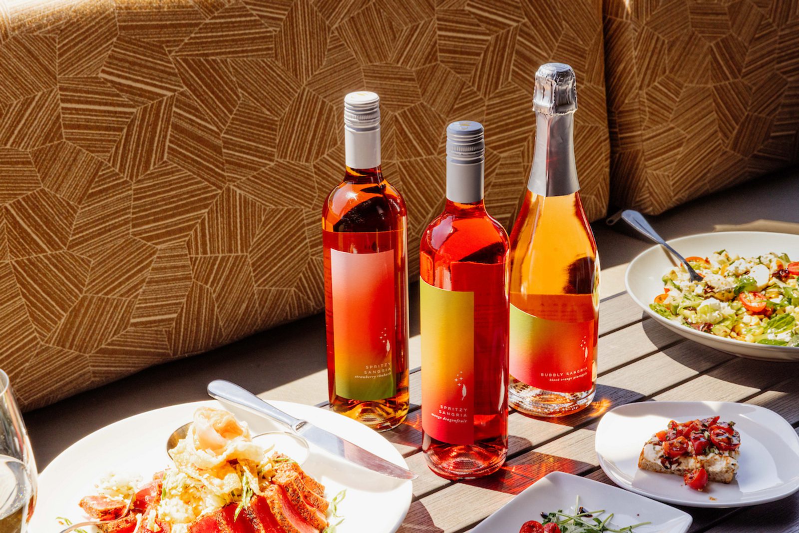 Three bottles of rosé on a sunlit wooden patio table with plates of salad and small appetizers