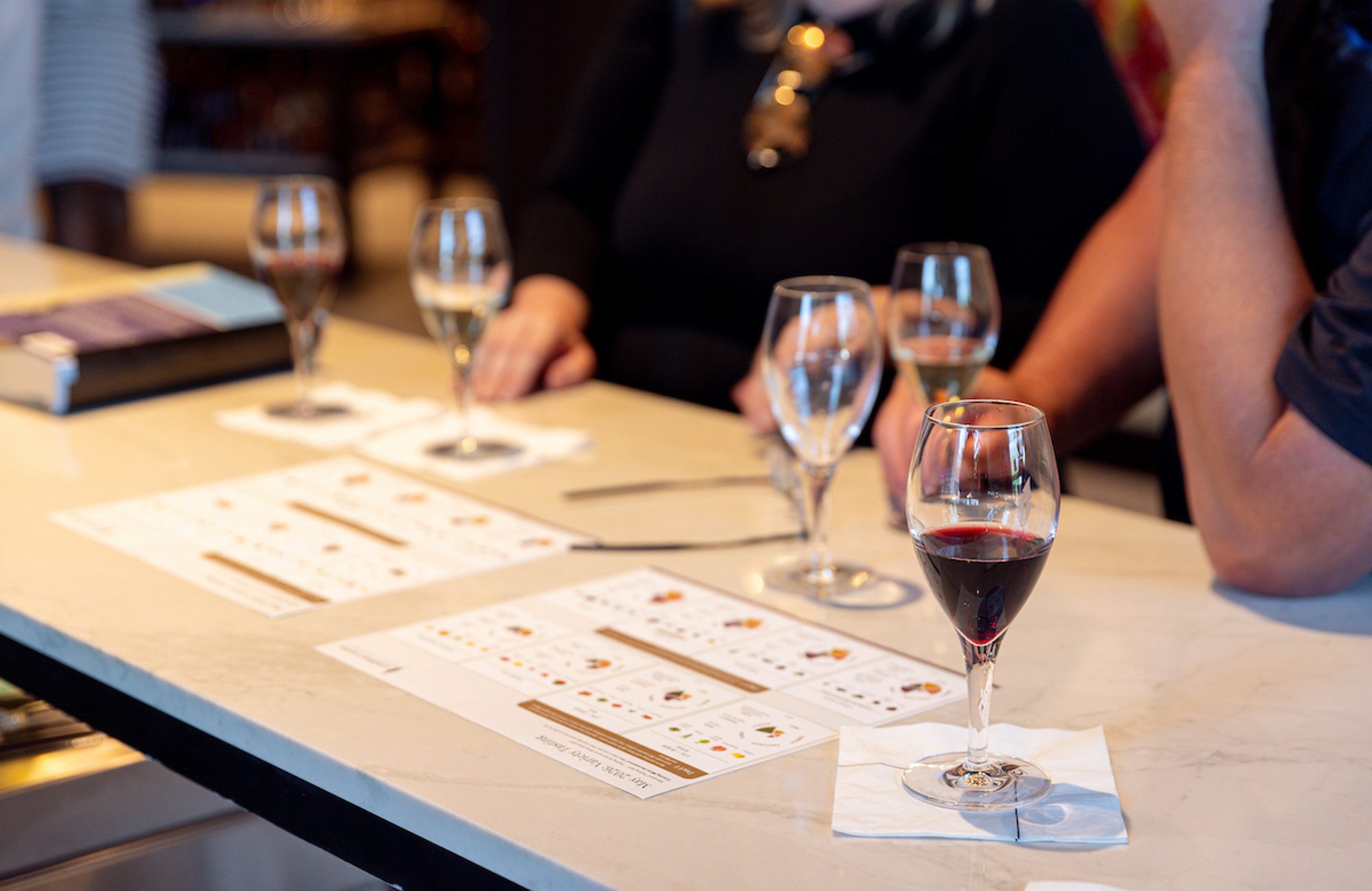 Wine tasting setup on a bar table with several glasses of red and white wine, tasting sheets, and people seated around the counter