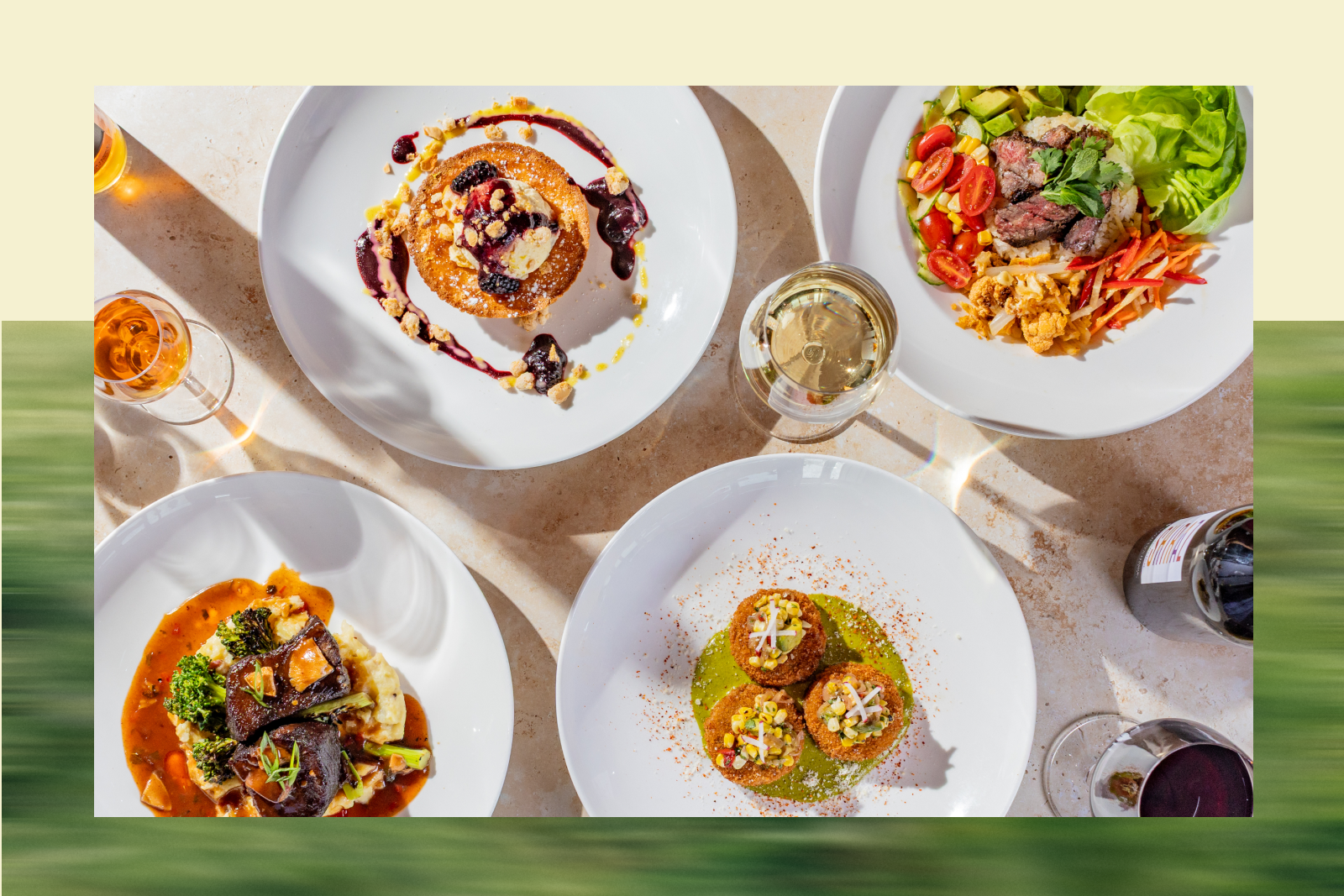 Overhead view of five white plates with assorted plated dishes and two glasses of wine on a marble table.