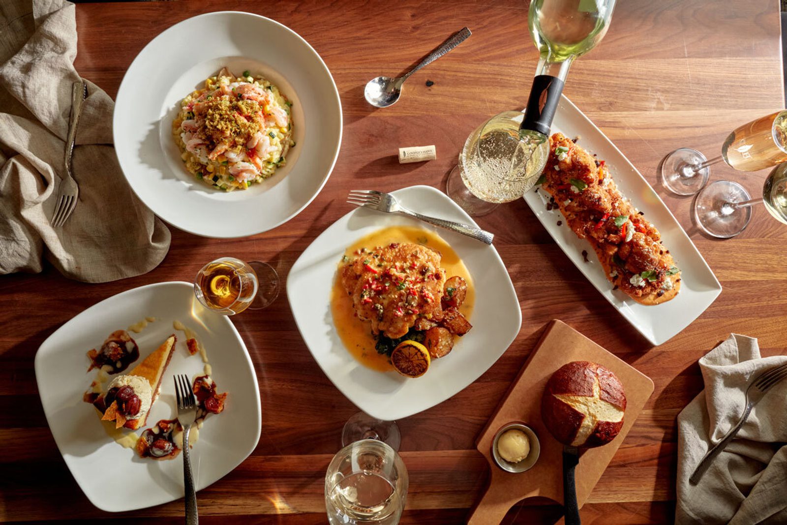 Overhead view of a wooden table set with plated gourmet dishes, bread, wine glasses, and silverware, with warm sunlight casting across the meal