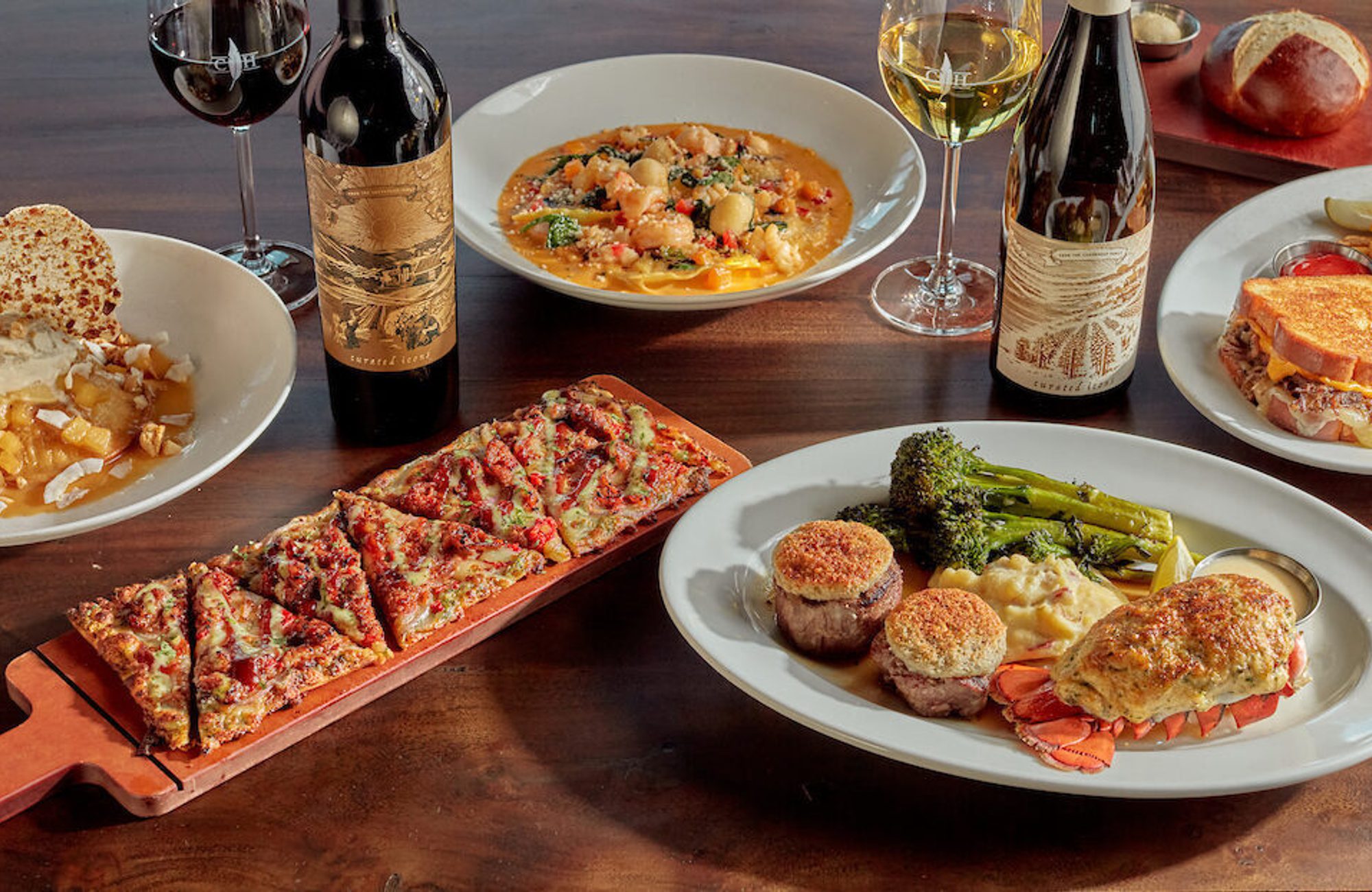 Table of gourmet dishes with wine glasses and bottles, including crab cake, flatbread, pasta, sandwich, fries, roasted vegetables, and dessert