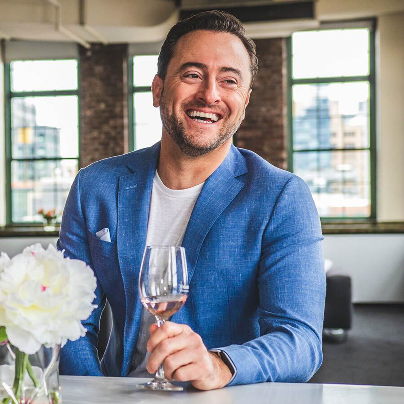 Man in blue blazer smiling while holding a wine glass at a table in a bright loft-style room