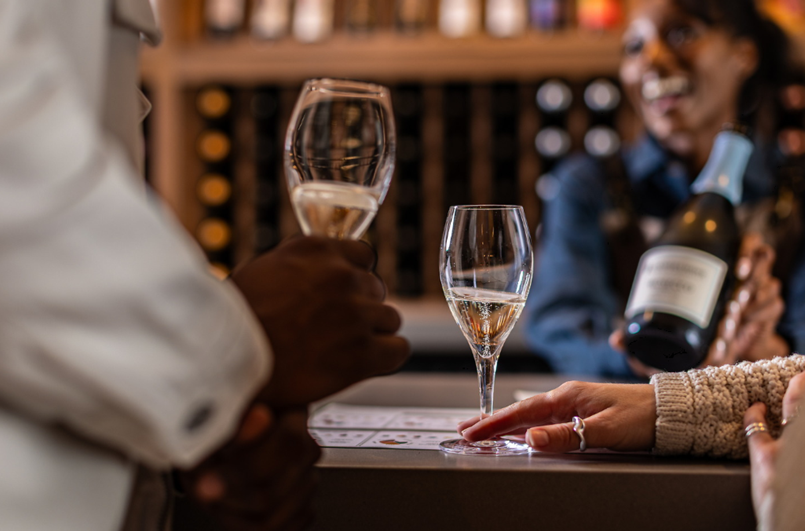 People at a wine tasting raise champagne flutes beside a bottle, with a glass on the table and a softly lit wine cellar in the background