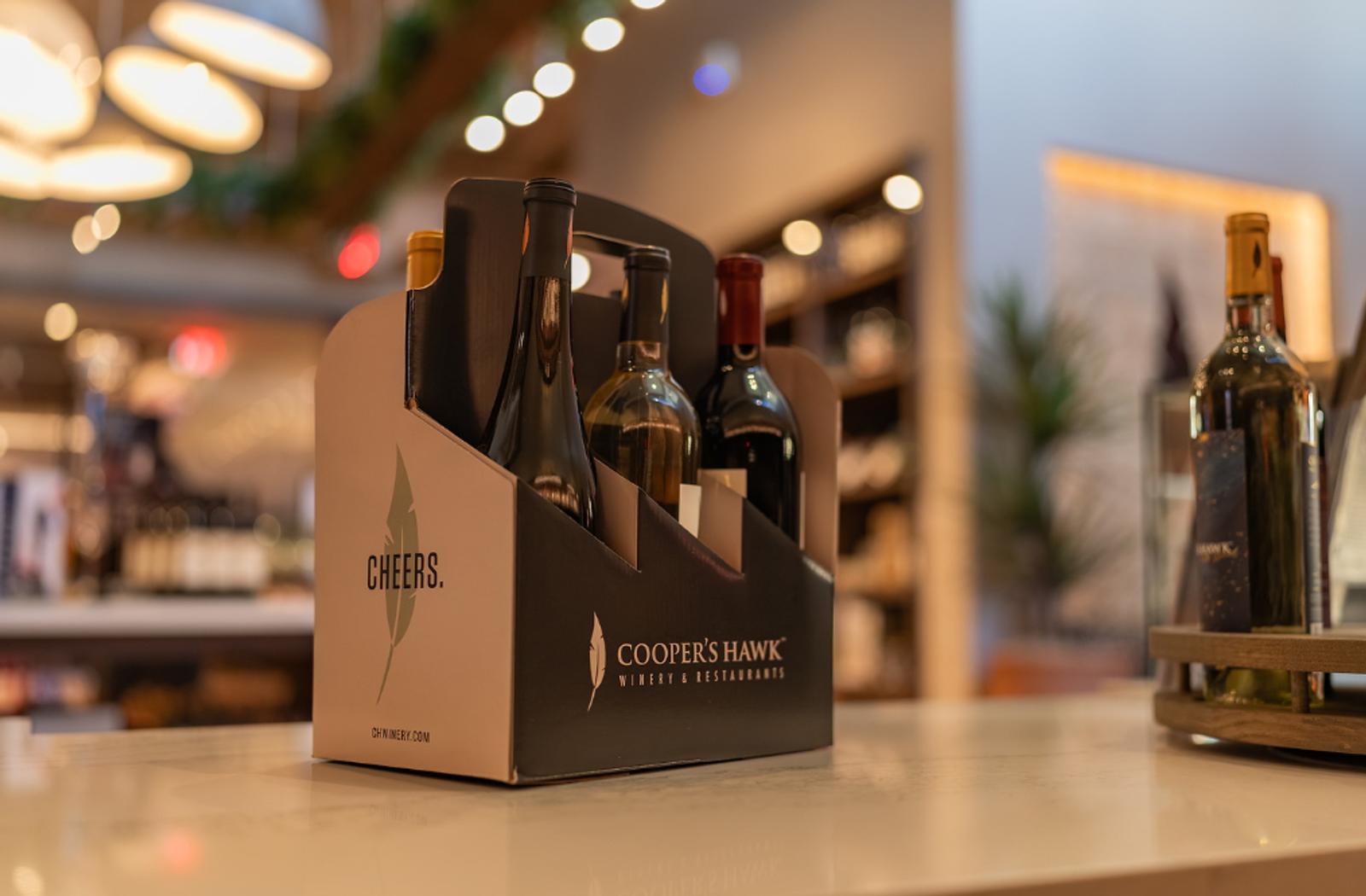 Wine bottles in a Cooper’s Hawk carrier on a countertop, with a softly blurred restaurant or wine shop interior and warm lights in the background