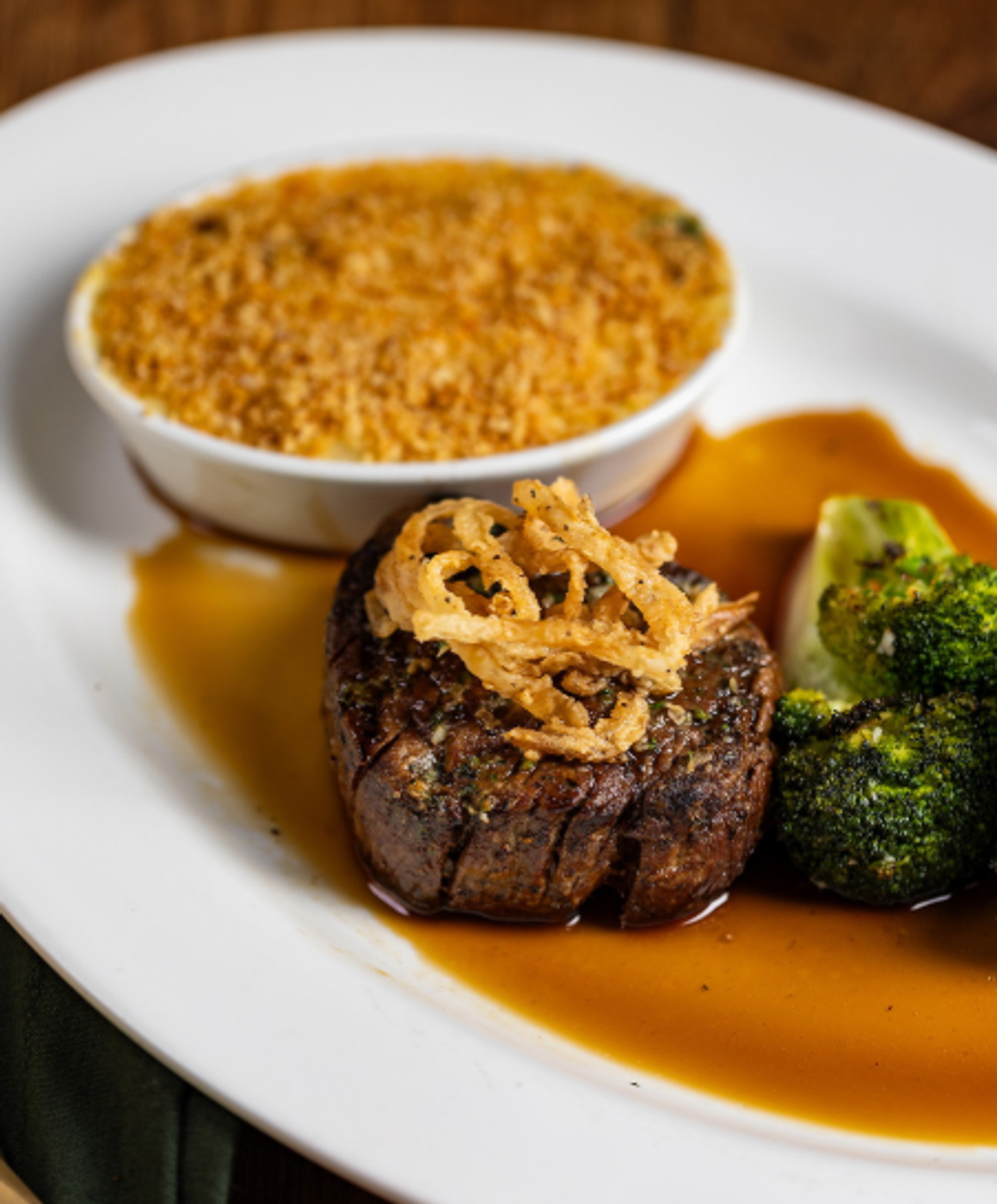 Plated filet steak topped with crispy fried onions, served with broccoli, brown sauce, and a ramekin of golden breadcrumb topping on a white plate