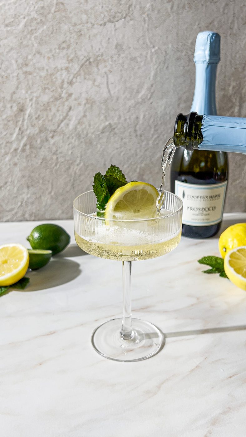 Coupe glass being filled with prosecco, garnished with lemon slice and mint; prosecco bottle and citrus halves on table.